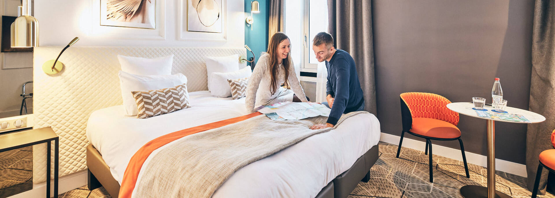 Pareja hablando frente a un plano sentada en la cama de una habitación de hotel