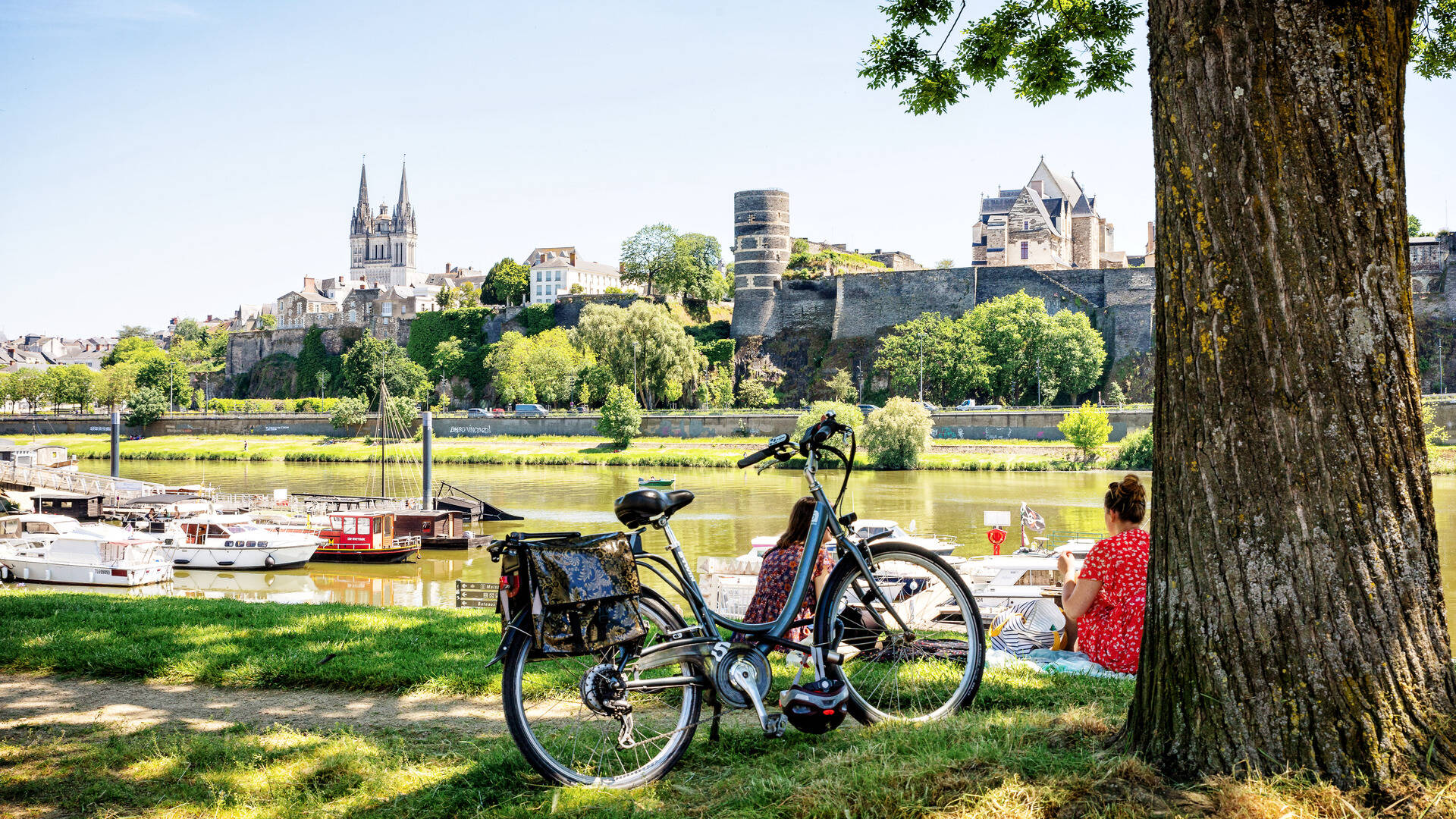 Cale de la Savatte, jeunes femmes assises sur l'herbe devant la Maine et le Château d'Angers, un vélo devant elles